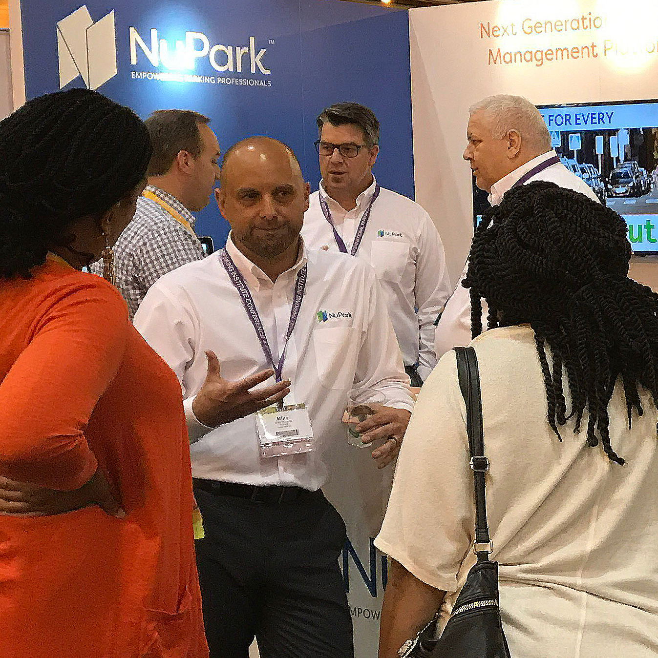 man in white shirt and black pants speaking to two ladies in white and orange clothes with men in the background talking with NuPark sign in the. back